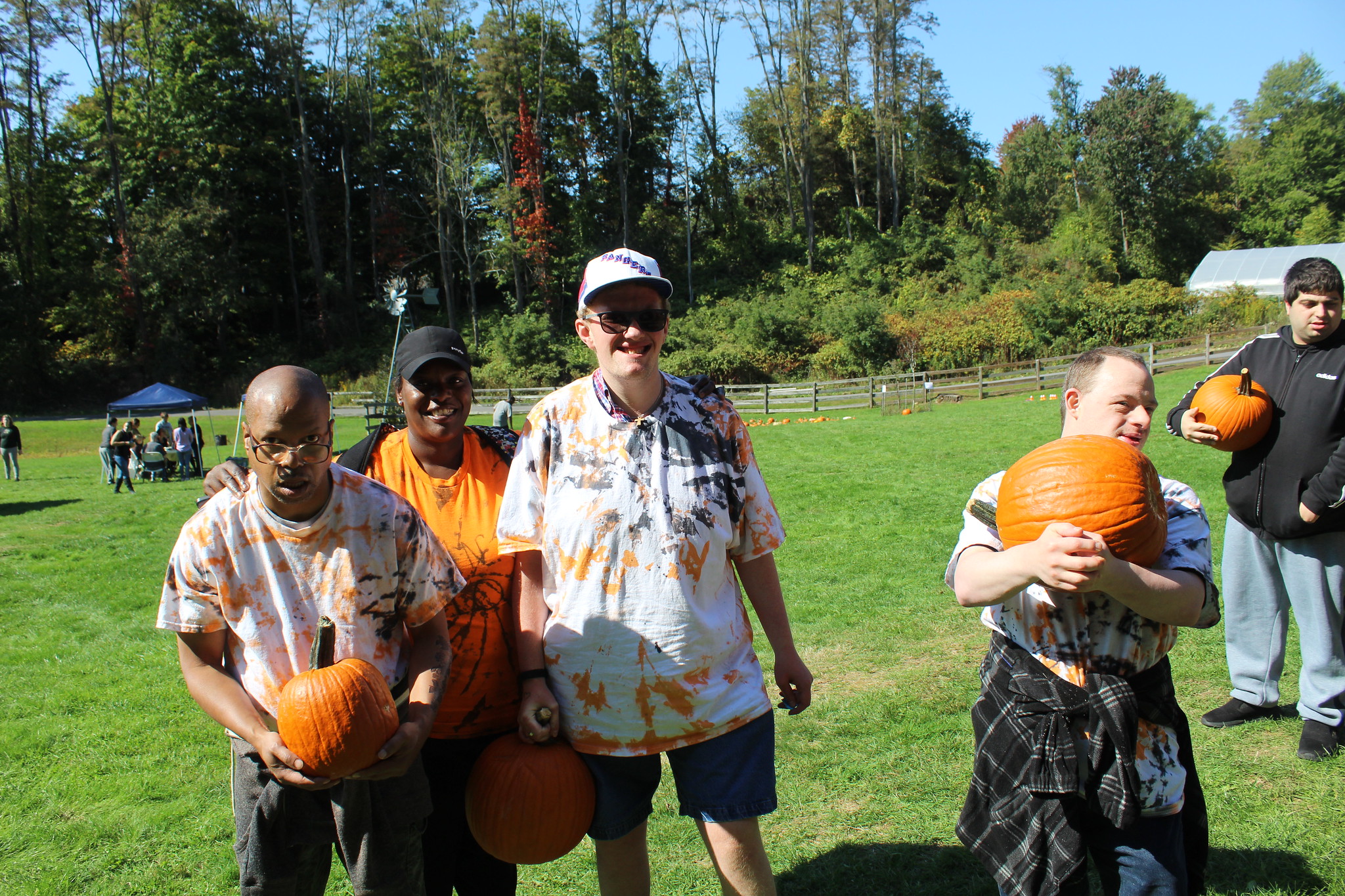 A group of three men in black and orange tie-dyed shirts hold large orange pumpkins. Their direct care person stands with them out on a field during the Cultivating Dreams Fall Festival at the Farm.