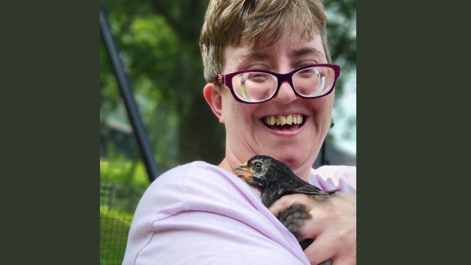 A women in short hair, red glasses and lavender shirt smiles as she holds a chicken that is a few weeks old.